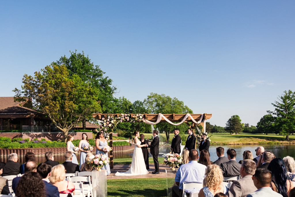 Outdoor wedding ceremony by a pond at Kemper Lakes Golf Club, with bride and groom at the altar, guests seated, and clear blue sky