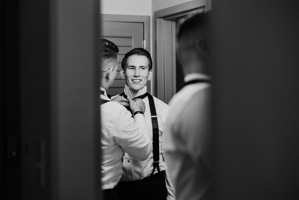 Black and white photo of groom smiling as another man helps him adjust his bow tie in front of a mirror while getting ready at Kemper Lakes Golf Club