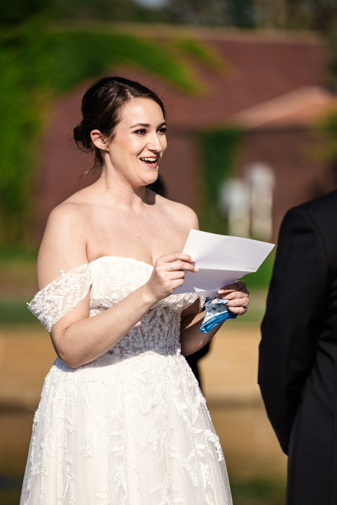 Bride in a white dress smiles while reading her vows during outdoor wedding ceremony at Kemper Lakes Golf Club