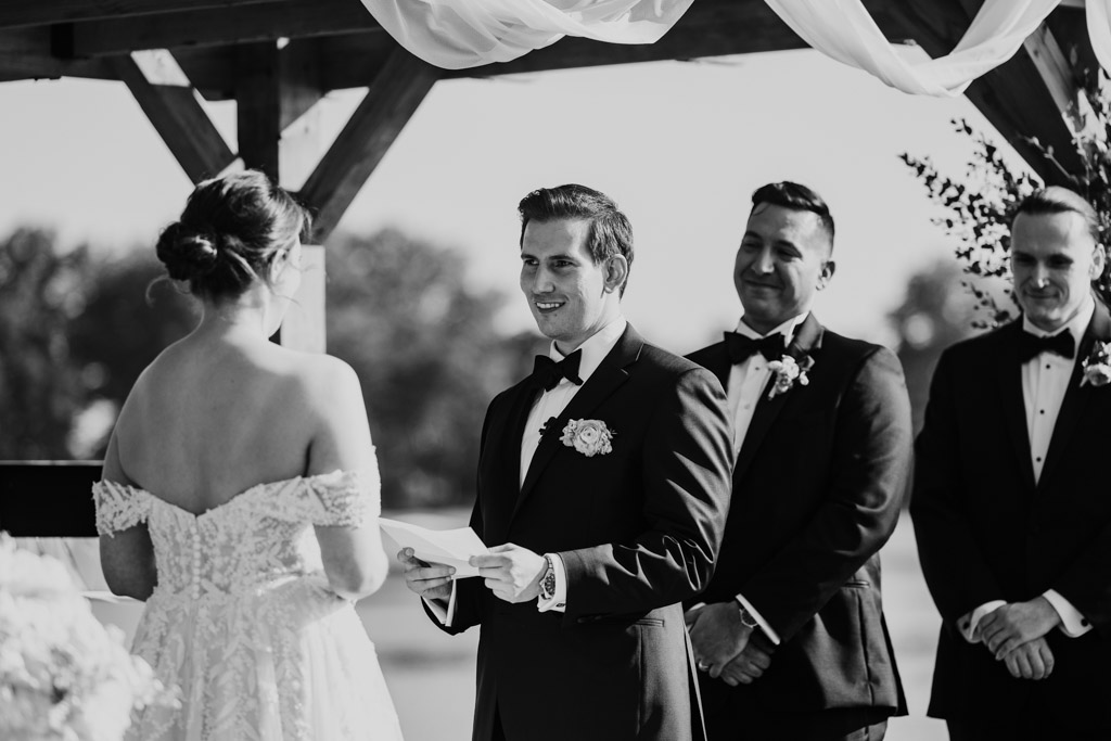 Black and white photo of groom reading vows to his bride at their outdoor Kemper Lakes Golf Club wedding ceremony, with groomsmen smiling behind him