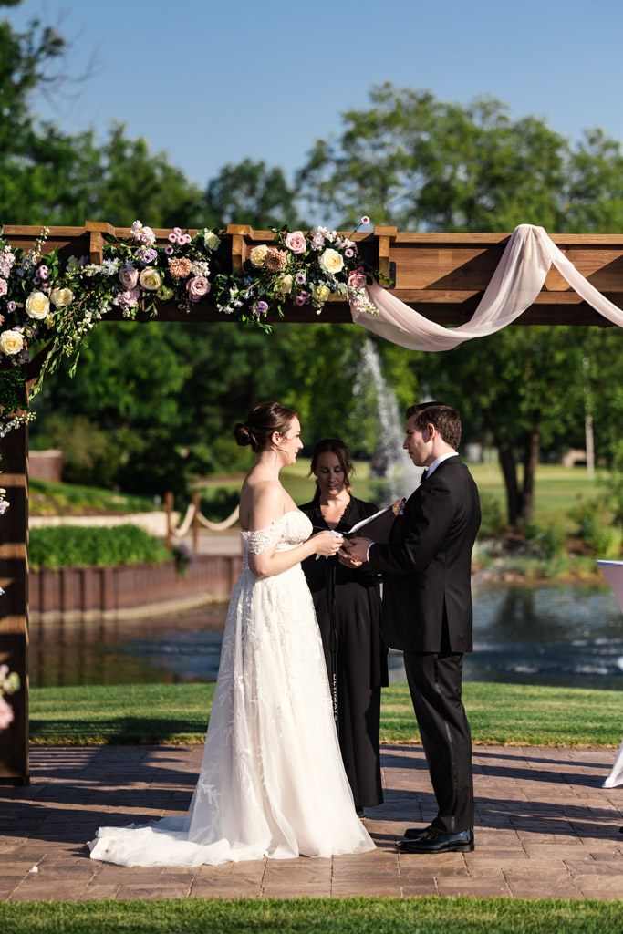 Bride and groom stand facing each other at an outdoor wedding ceremony under a flower-decorated wooden arch at Kemper Lakes Golf Club