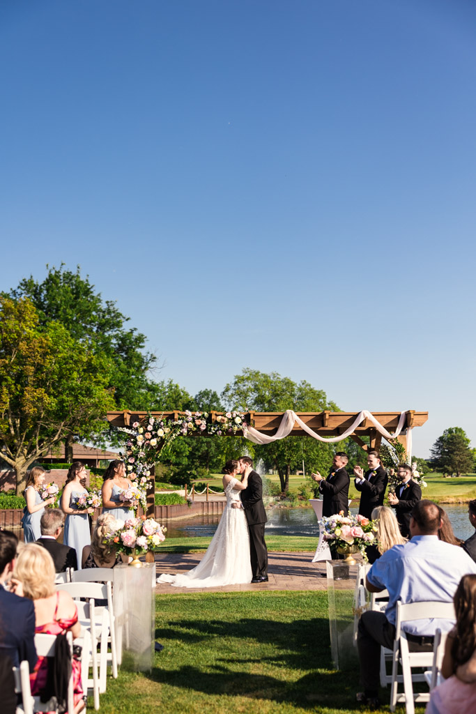 Bride and groom kiss under a floral arch at their sunny wedding ceremony at Kemper Lakes Golf Club