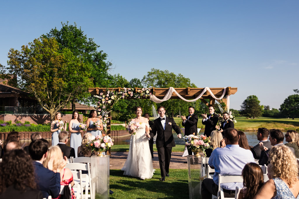 Happy newlyweds walk down the aisle outdoors after their wedding ceremony at Kemper Lakes Golf Club, with wedding party and guests celebrating