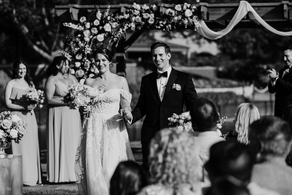 Black and white photo of bride and groom walking down the aisle, smiling, after their outdoor wedding ceremony at Kemper Lakes Golf Club, with guests watching