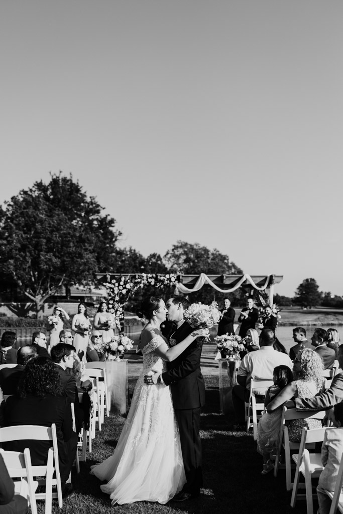 Black and white photo of bride and groom kissing mid-aisle at their outdoor wedding ceremony at Kemper Lakes Golf Club, surrounded by seated guests