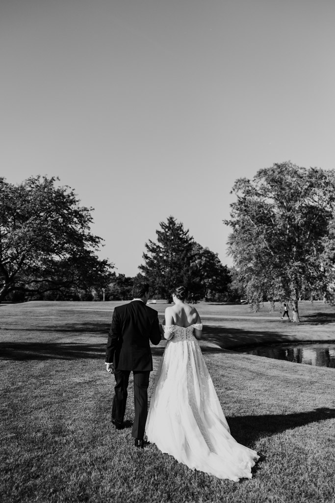 Black and white photo of bride and groom walking hand in hand across a grassy field under a clear sky at Kemper Lakes Golf Club