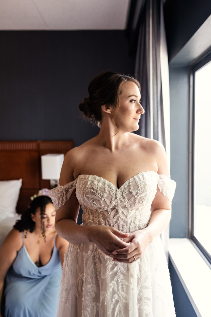 A bride in a white gown stands by a window while getting ready before her wedding at Kemper Lakes Golf Club, while a bridesmaid sits behind her on a bed