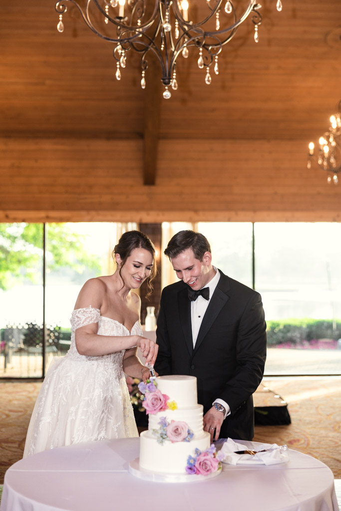 Bride and groom smile as they cut their wedding cake decorated with pink and purple flowers at Kemper Lakes Golf Club