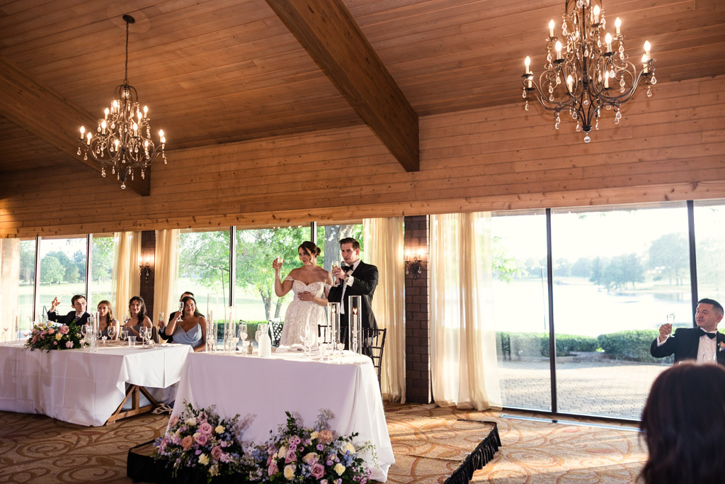 Bride and groom give a toast at their wedding reception in a wood-paneled room with large windows and chandeliers at Kemper Lakes Golf Club