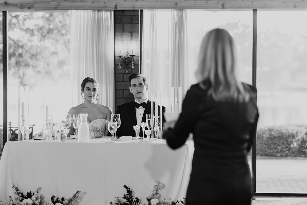 Black and white photo of bride and groom sitting at the head table, listening to groom's mother give a speech during their Kemper Lakes Golf Club wedding reception
