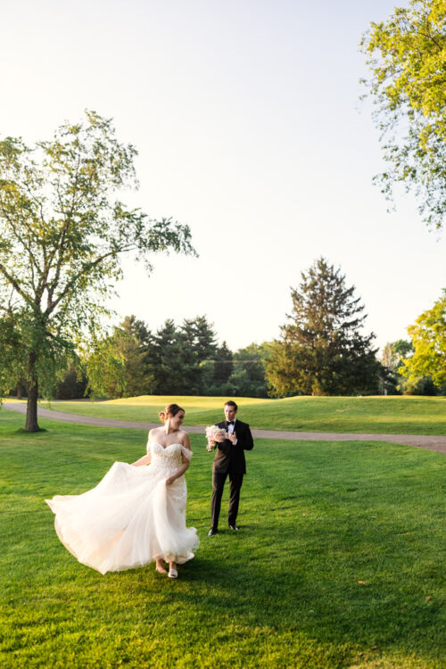 Bride twirls her dress while walking with the groom on a sunny, grassy lawn at Kemper Lakes Golf Club, with trees in the background