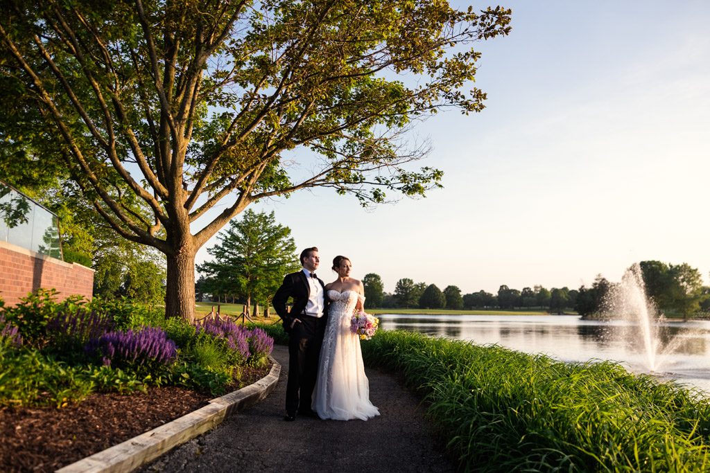 Bride and groom standing on a path by a lake with a fountain at Kemper Lakes Golf Club, surrounded by greenery and flowers