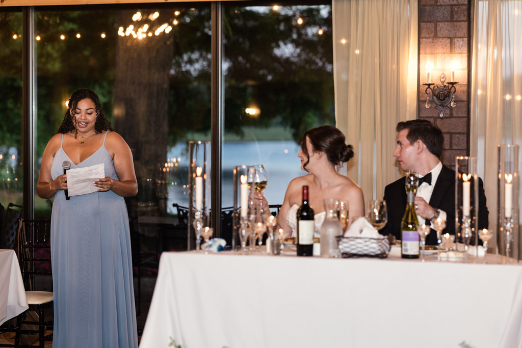 Maid of Honor in a blue dress gives a speech during wedding reception at Kemper Lakes Golf Club as the bride and groom listen from their table