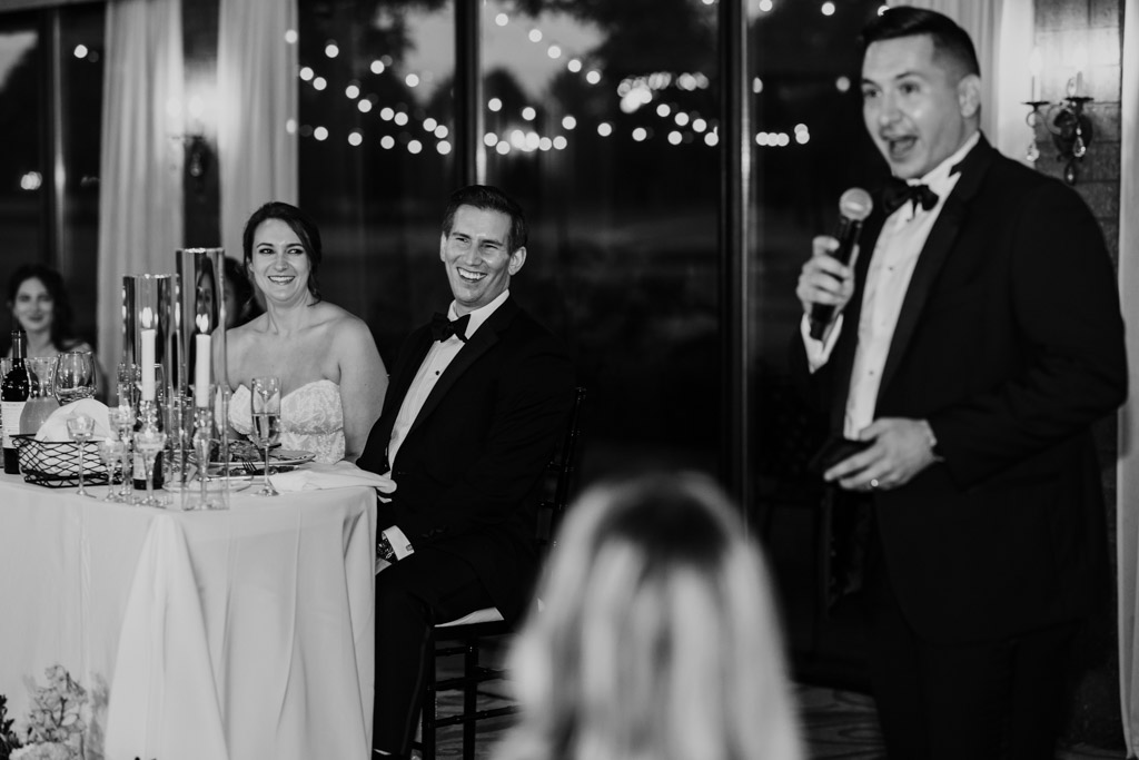 Black and white photo of newlyweds smiling while Best Man gives speech during wedding reception at Kemper Lakes Golf Club