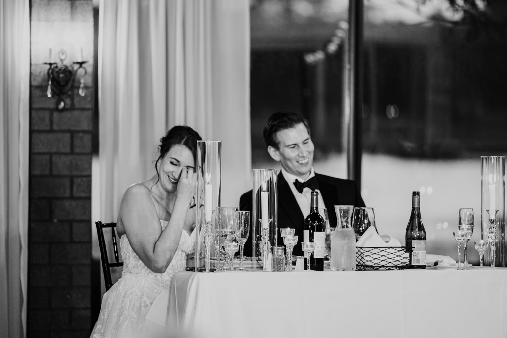 Black and white photo of bride and groom laughing seated at their table during wedding reception at Kemper Lakes Golf Club