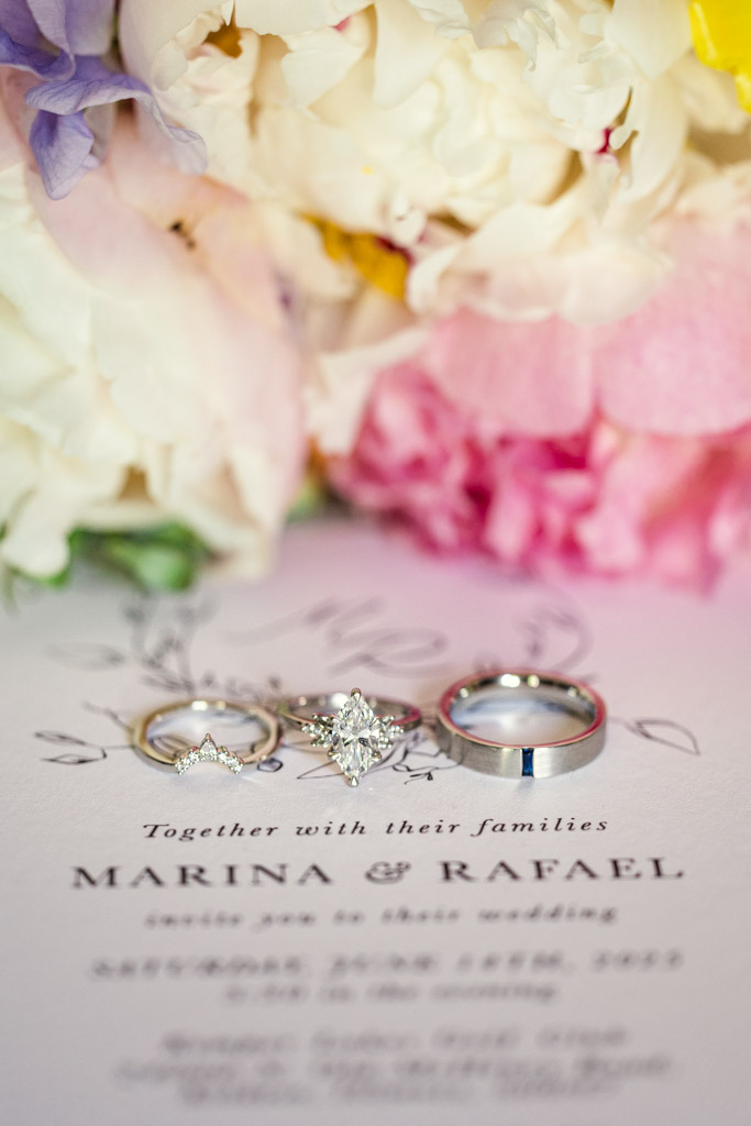 Three wedding rings rest on a wedding invitation, with colorful flowers in the background for Kemper Lakes Golf Club wedding celebration