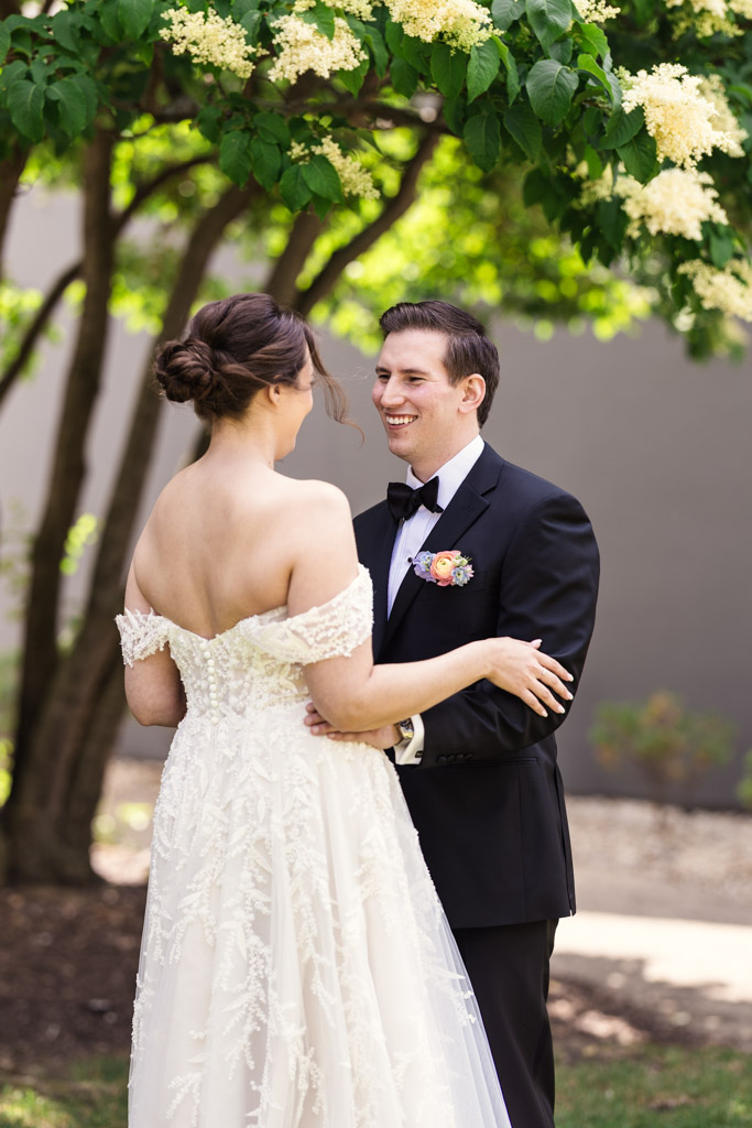 Bride and groom smile at each other outdoors under a flowering tree during their first look before their wedding at Kemper Lakes Golf Club
