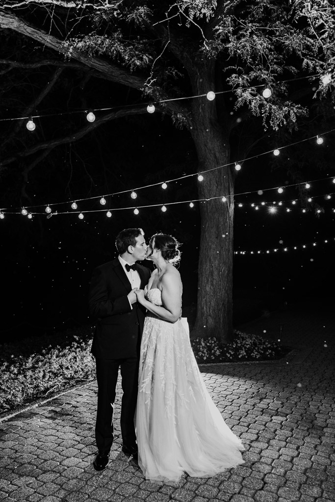 Black and white photo of bride and groom kissing under string lights at night on a stone path at Kemper Lakes Golf Club