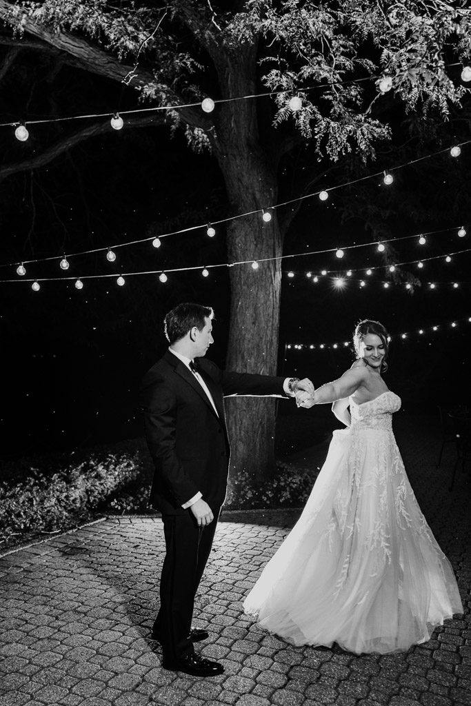 Black and white photo of bride and groom dancing together outdoors at night under string lights at Kemper Lakes Golf Club