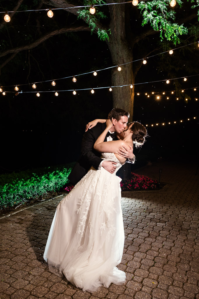 Bride and groom kiss under string lights at night outside Kemper Lakes Golf Club