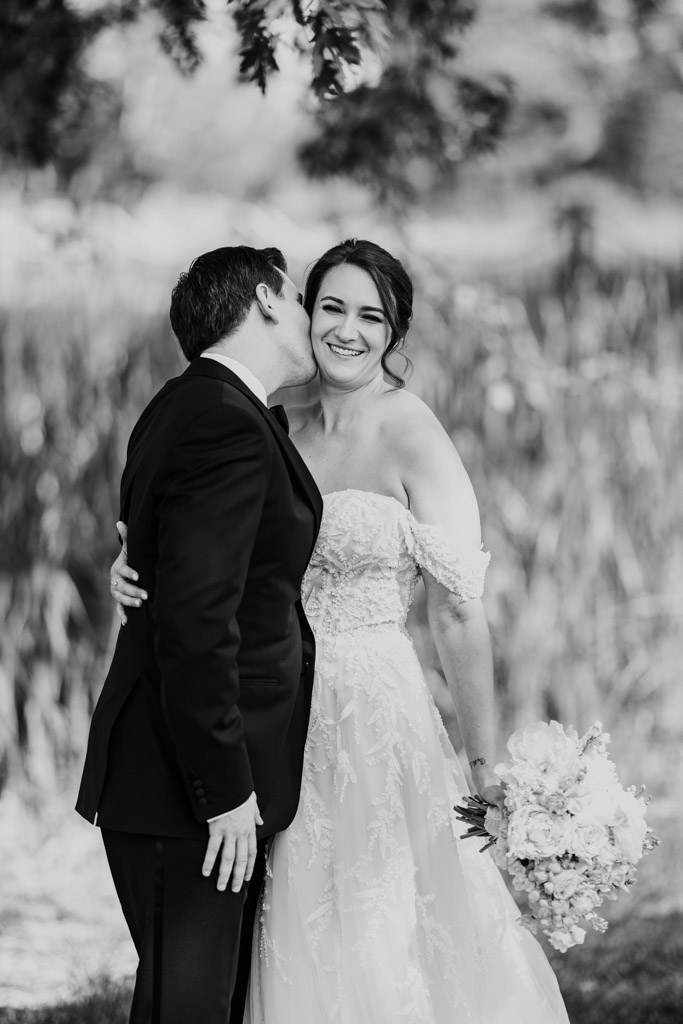 Black and white photo of bride smiling as groom kisses her cheek outdoors at Kemper Lakes Golf Club, both in wedding attire, bouquet in her hand