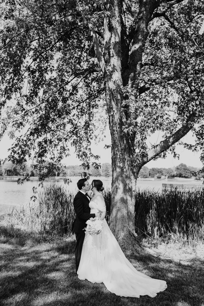 Black and white photo of bride and groom kiss under a large tree by a pond at Kemper Lakes Golf Club on their wedding day