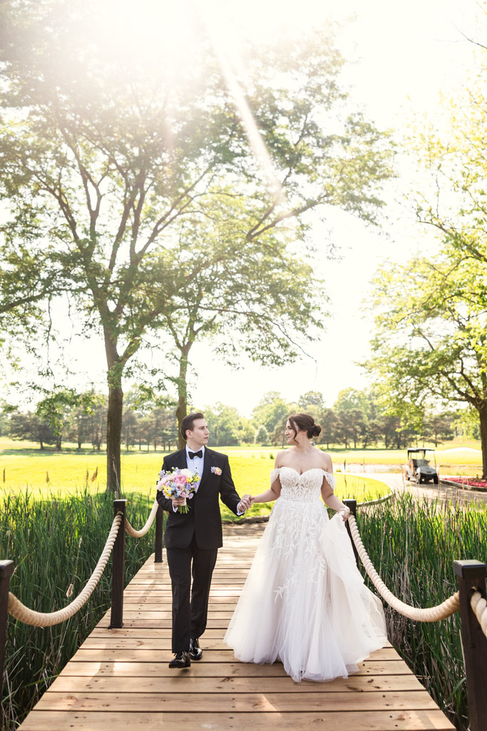 Bride and groom walk hand in hand on a sunlit wooden bridge surrounded by trees and greenery at Kemper Lakes Golf Club