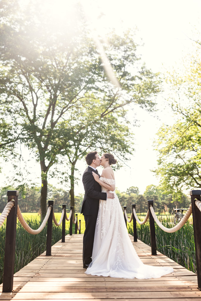 Bride and groom kiss on a wooden bridge under sunlight, surrounded by green trees at Kemper Lakes Golf Club