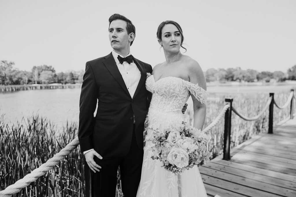 Black and white portrait of bride and groom in formal attire standing together on a wooden dock by a pond at Kemper Lakes Golf Club, gazing into the distance