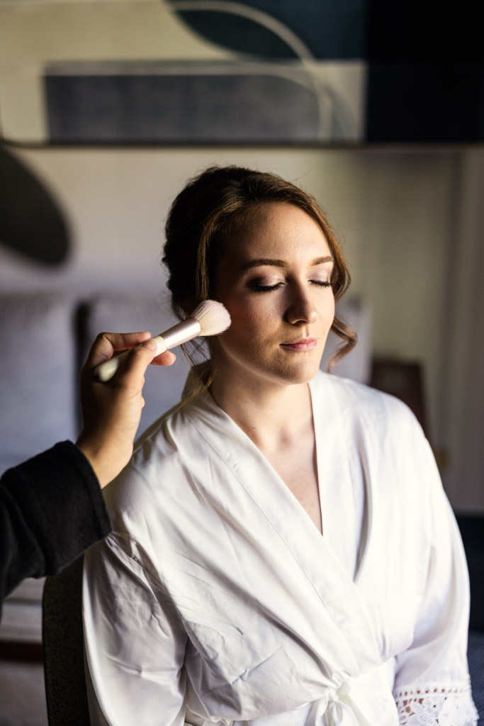 Bride in a white robe has her makeup done with a brush while her eyes are closed as she gets ready before her wedding at Kemper Lakes Golf Club