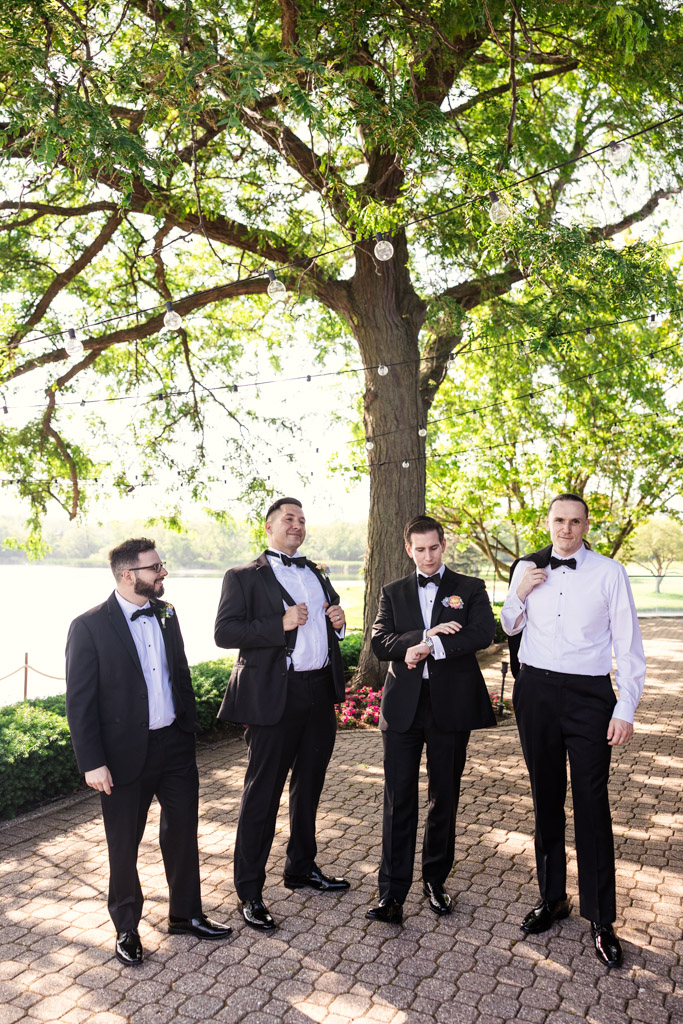 Groom and groomsmen in tuxedos stand outdoors under a large tree at Kemper Lakes Golf Club, smiling and adjusting their jackets