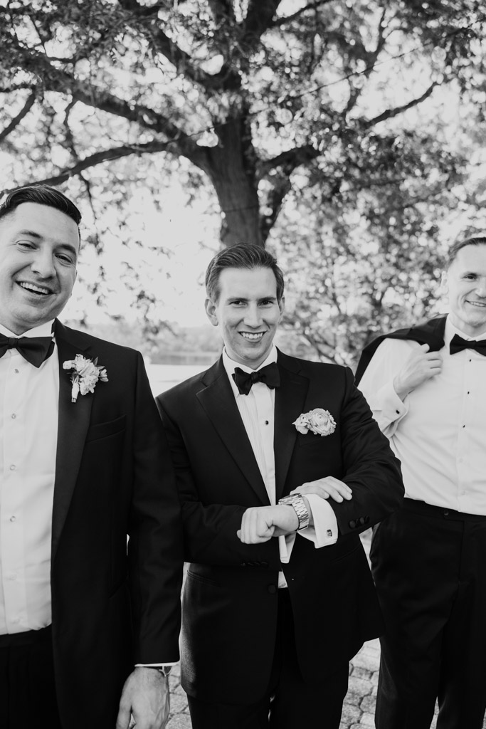 Black and white photo of groom in tuxedo showing off his watch and cufflinks at Kemper Lakes Golf Club, with trees in the background