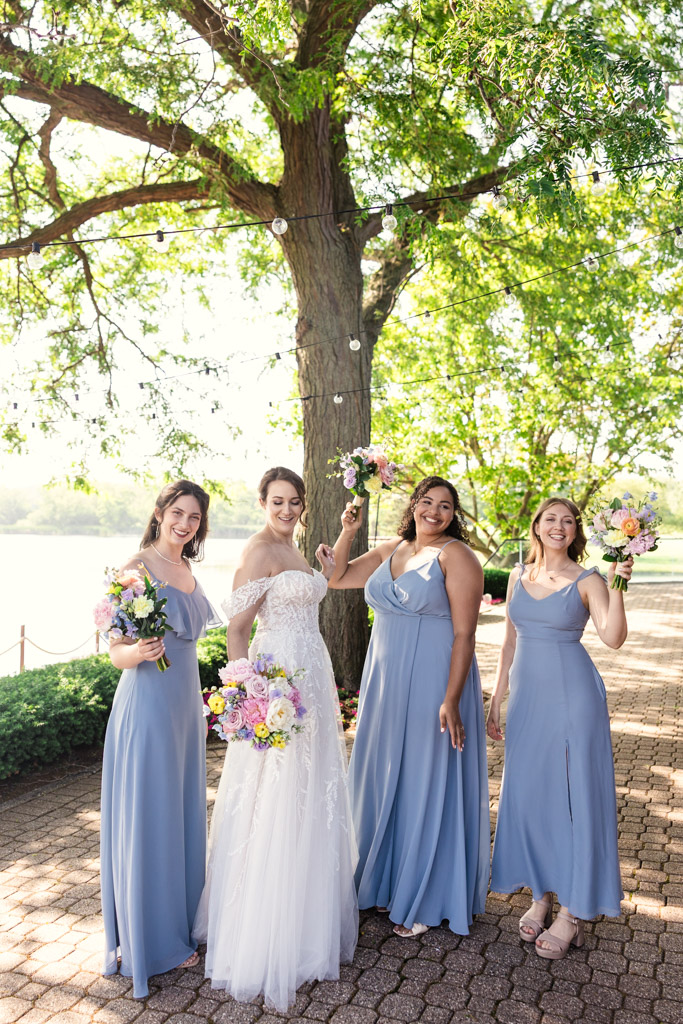 Bride in a white dress and bridesmaids in light blue dresses pose with bouquets under a large tree at Kemper Lakes Golf Club