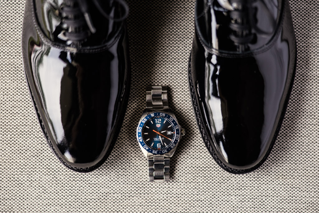 Groom's shoes and stainless steel wristwatch with a black and blue face rest on a textured surface before wedding at Kemper Lakes Golf Club