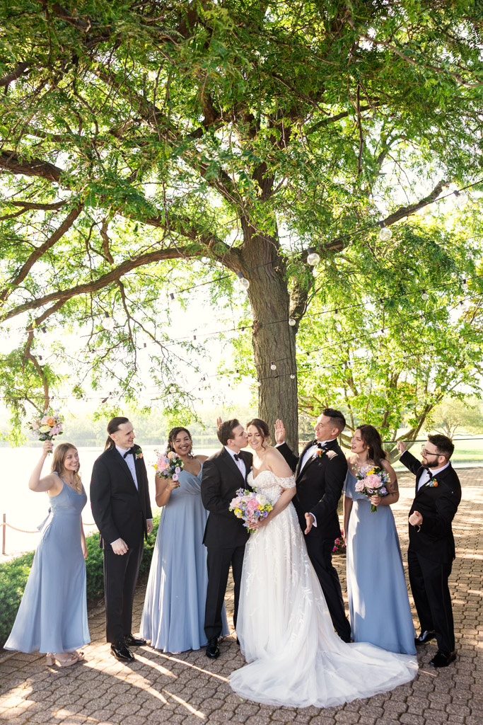 Joyful wedding party poses under a large tree at Kemper Lakes Golf Club, with bridesmaids in blue dresses and groomsmen in tuxedos