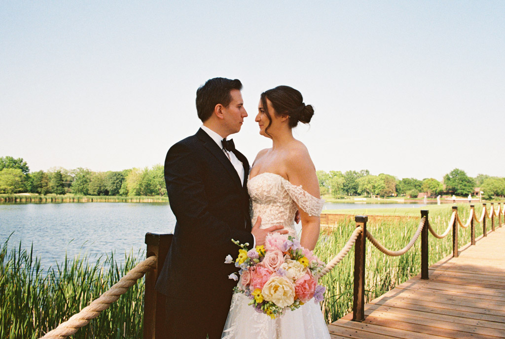 Film photo of bride and groom standing on a dock by Kemper Lakes Golf Club, gazing at each other and smiling on their wedding day