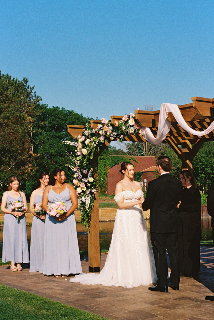 Film photo of bride and groom exchanging vows outdoors under a floral arch at Kemper Lakes Golf Club, with bridesmaids nearby