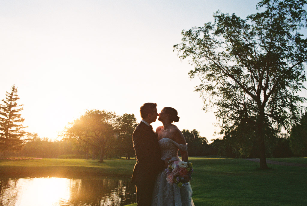 Film photo of bride and groom embracing at sunset on the grounds of Kemper Lakes Golf Club, with trees and a pond in the background