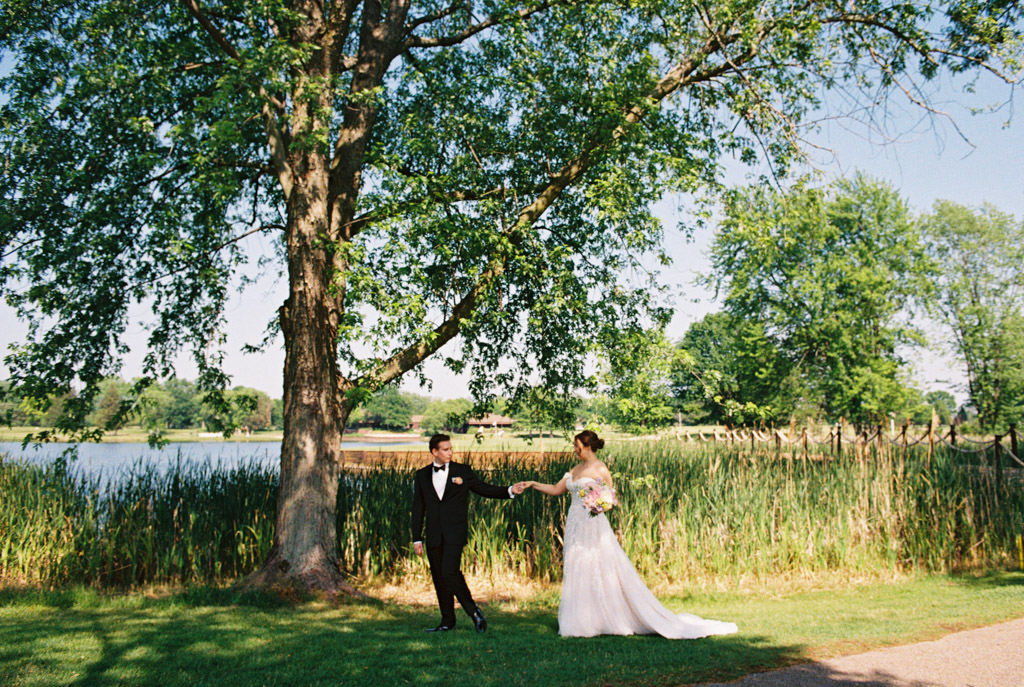 Newlyweds walks hand in hand under a large tree by a pond on a sunny day at Kemper Lakes Golf Club