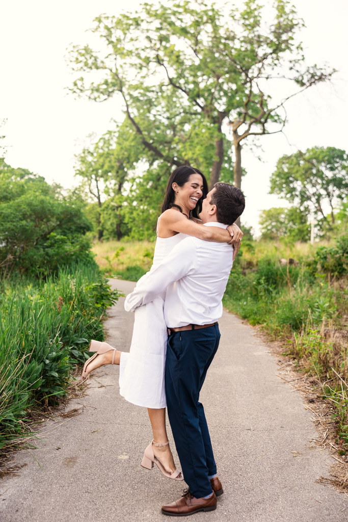 A man lifts his partner wearing a white dress as they stand on a path surrounded by greenery in Lincoln Park during spring engagement session