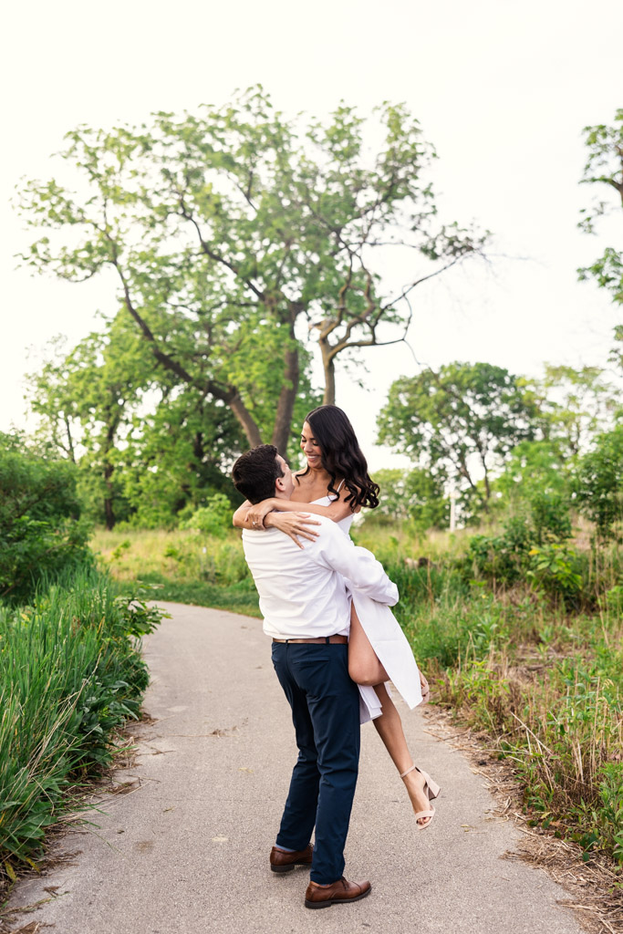 A man lifts his partner in his arms on a path surrounded by greenery and trees in Lincoln Park during spring engagement session