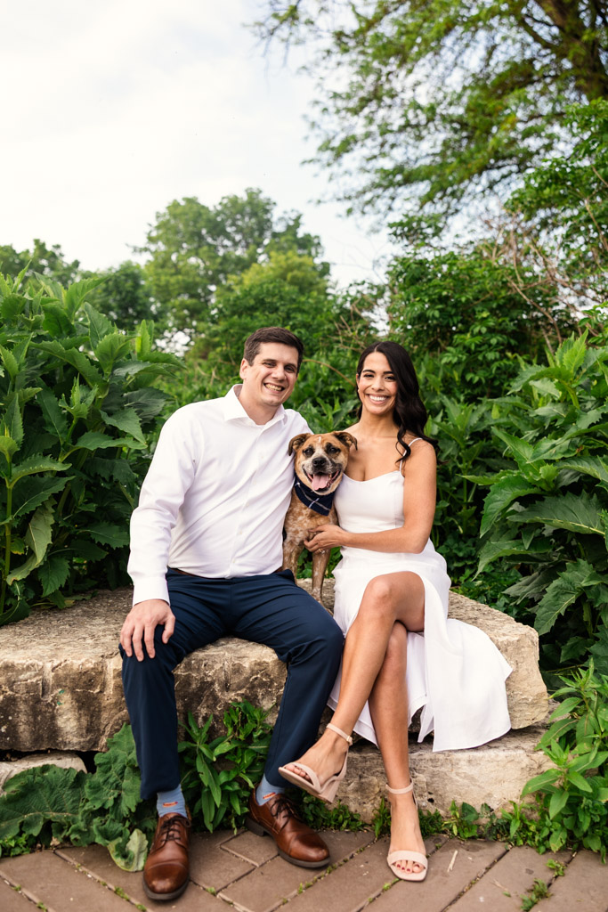 A smiling couple sits on a stone bench with their happy dog, surrounded by lush greenery during Lincoln Park engagement session