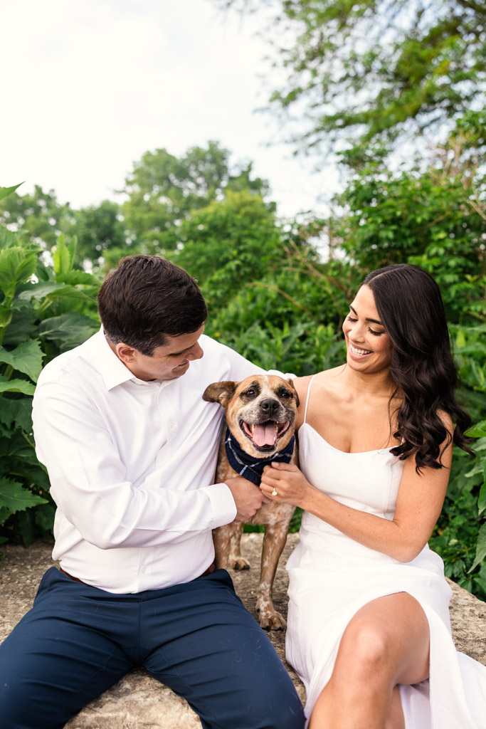 Engaged couple sits outdoors with their happy dog between them, surrounded by greenery in Lincoln Park