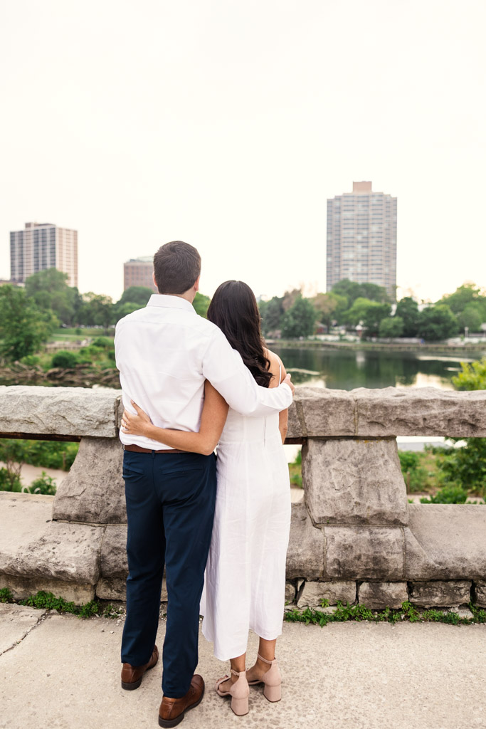 Engaged couple holds each other standing on a stone bridge in Lincoln Park, gazing at downtown Chicago