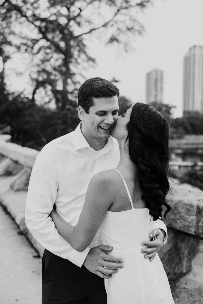 Black and white photo of woman kissing her partner on the cheek as they embrace outdoors during Lincoln Park engagement session