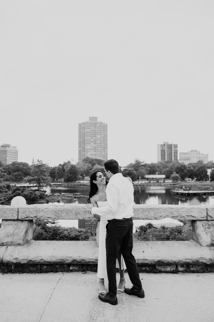 Black and white photo of couple smiling and dancing on a bridge in Lincoln Park with Chicago buildings in the background during their late spring engagement session