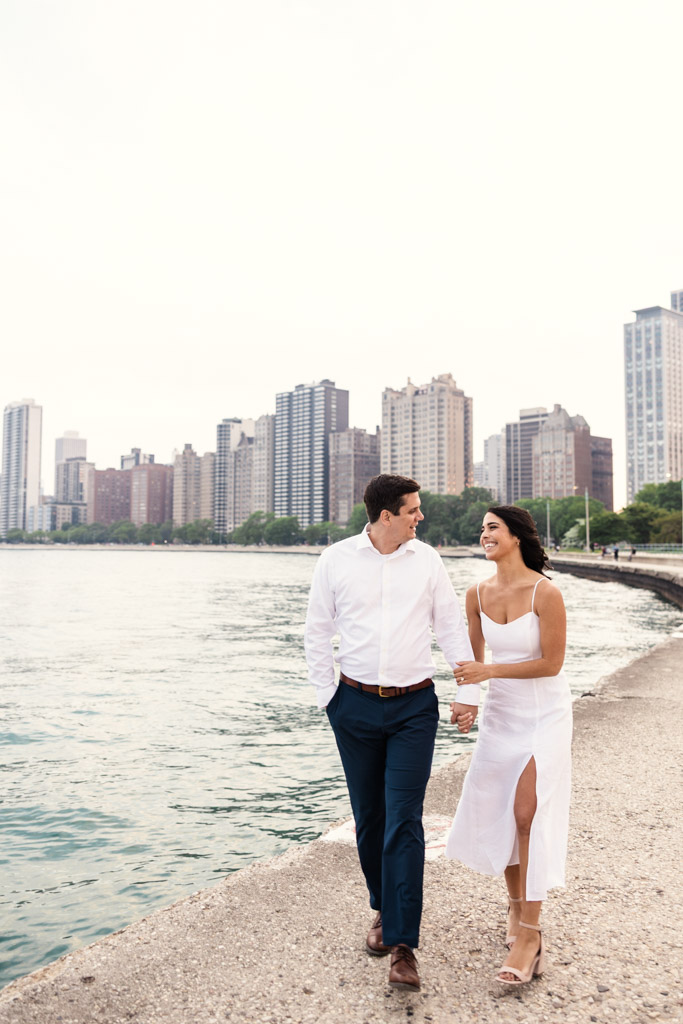 Happy engaged couple walks arm in arm by Lake Michigan at North Avenue Beach in Lincoln Park with downtown Chicago buildings in the background