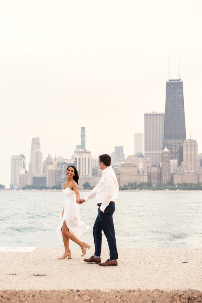 Couple holds hands walking by the water at North Avenue Beach with the Chicago skyline in the background during Lincoln Park engagement session