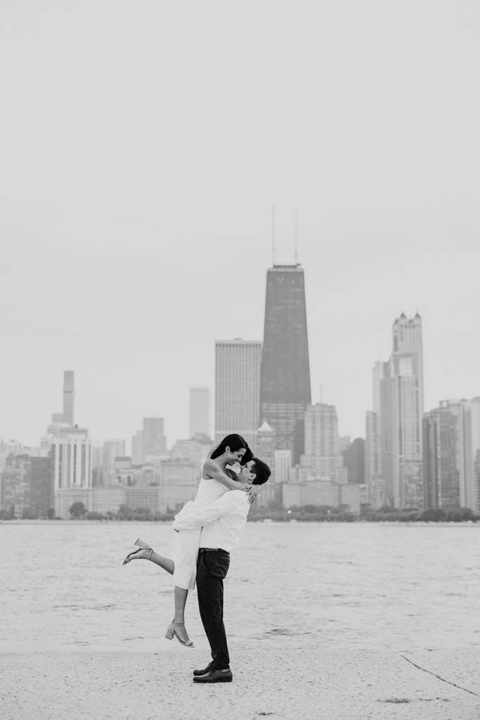 Black and white photo of man lifting his partner as they walk along the shore of Lake Michigan during Lincoln Park engagement session