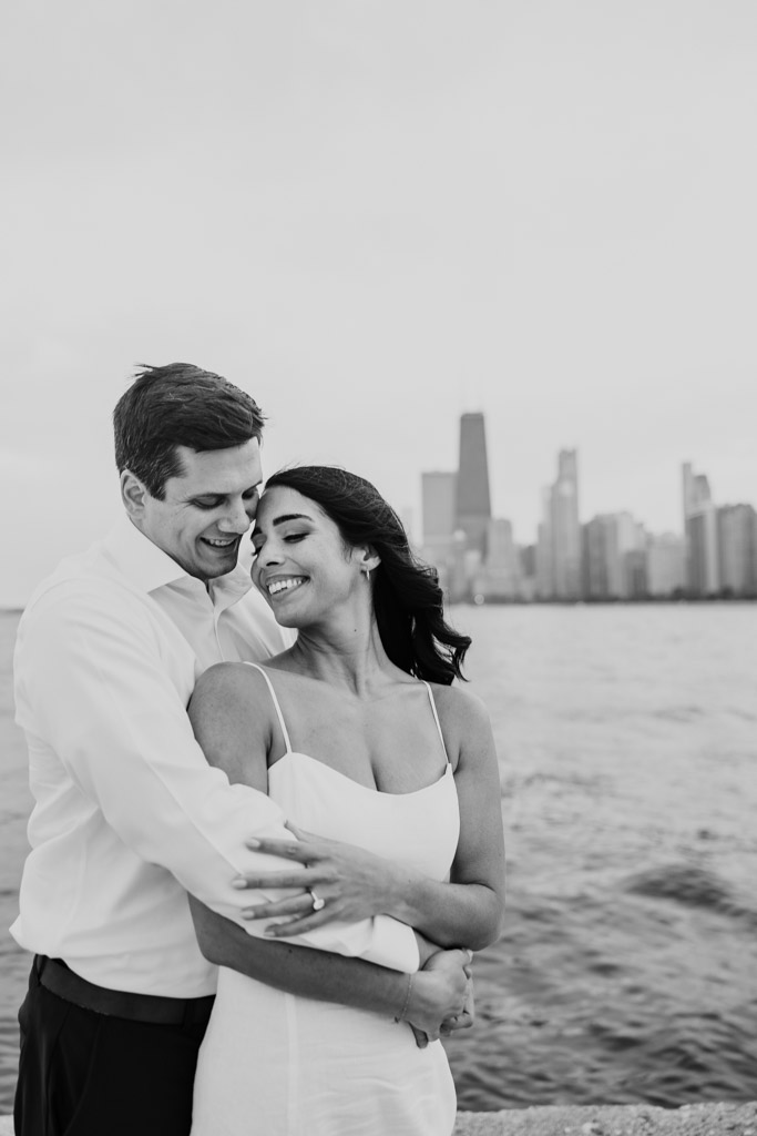 Black and white photo of engaged couple smiling and holding each other by the waterat North Avenue Beach with the Chicago skyline behind them